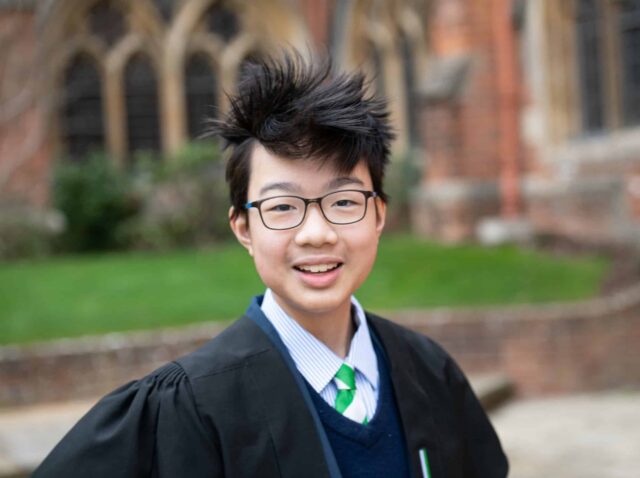 Boy in gown and tie smiling at the camera in the grounds of Radley College
